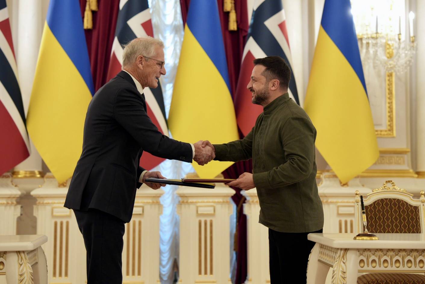 Bilde av Støre og  Zelenskyj som håndhilser og utveksler en mappe. I bakgrunnen er norske og ukrainske flagg.