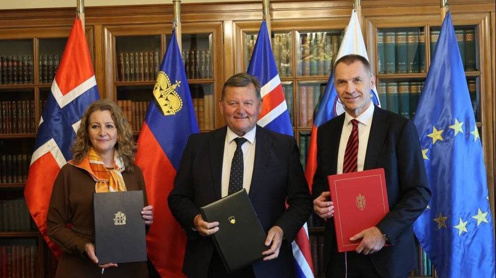 Tre personer står foran EU- og nasjonale flagg og holder dokumenter i et formelt rom med bokhyller. Nederst står