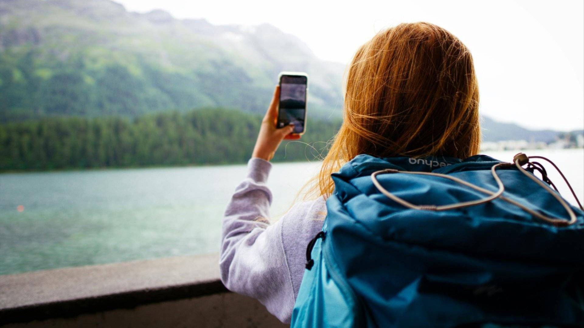 Bilde en jente som står med ryggen til kamera og ser mot et vann og fjell mens hun tar et bilde med mobiltelefonen
