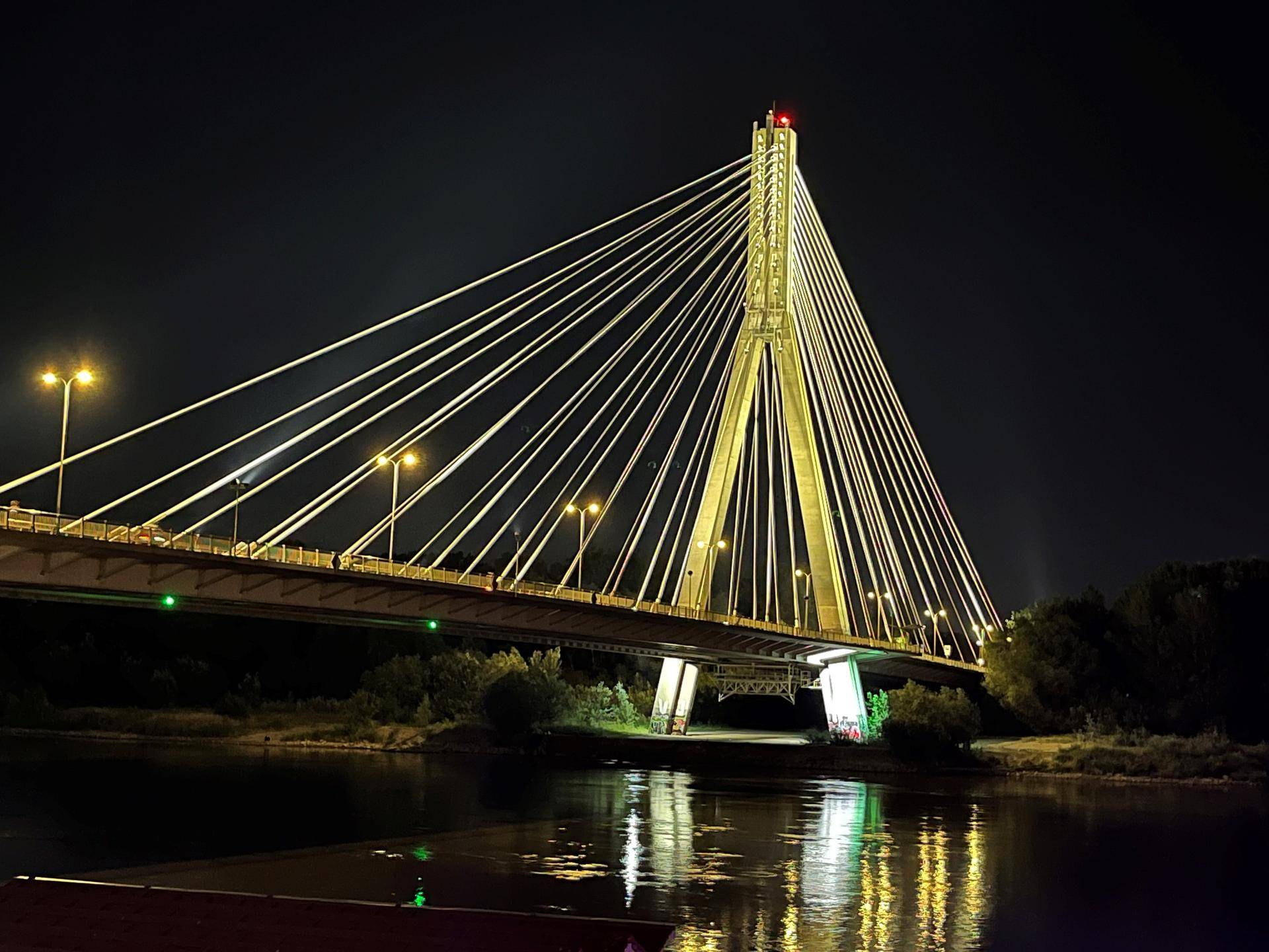 Foto av en bro opplyst om natten. Broen som befinner seg i Warzawz, Polen heter Most Świętokrzyski. På engels oversettes dette til Holy Cross Bridge. 
